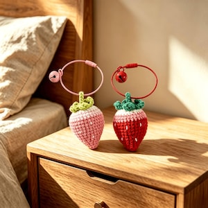 May include: Two crochet strawberry keychains with green tops, one in light pink and one in red. Each strawberry is attached to a circular metal wire ring featuring a small round bell. The light pink strawberry has a pink ring, and the red strawberry has a red ring.