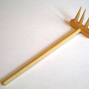 May include: A wooden hand rake with five tines. The rake is made of light brown wood and has a long handle.