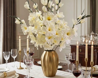May include: A large bouquet of white gladiolus flowers with yellow centers arranged in a matte gold, ribbed vase. The vase sits on a dining table set with white plates, gold napkin rings, crystal glassware, and tall taper candles in gold holders.