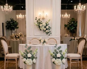 May include: A round sweetheart table covered with a white tablecloth, featuring two floral arrangements of white roses, hydrangeas, and eucalyptus leaves, flanked by two light-colored upholstered chairs in a room with chandeliers.