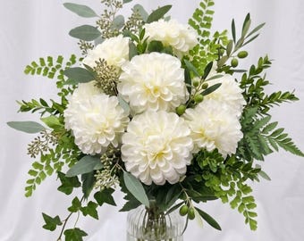 May include: A bouquet of white dahlia flowers arranged with green eucalyptus, fern fronds, ivy, and olive branches with small green olives. The floral arrangement is displayed in a clear, vertically ribbed glass vase.