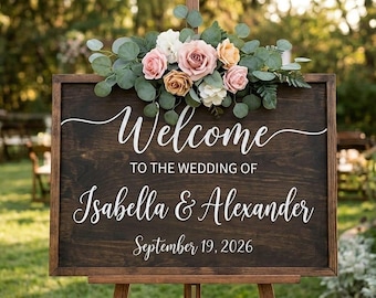 May include: A rectangular dark wood wedding sign with white lettering that reads Welcome TO THE WEDDING OF Isabella & Alexander September 19, 2026. The sign is decorated with a cluster of pink, peach, and white roses and green eucalyptus leaves.