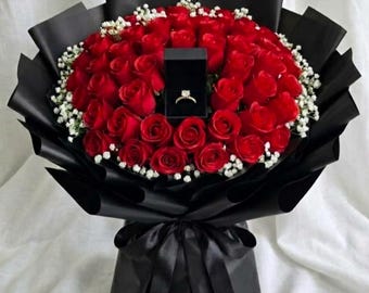 May include: A large bouquet of 72 red rose buds accented with small white baby's breath flowers, wrapped in black paper with a black ribbon bow. A small black square ring box containing a gold ring with a single stone sits in the center.