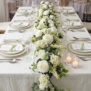 Puede incluir: Una mesa de comedor larga preparada para un evento formal con mantel blanco, centro de mesa floral blanco con rosas y vegetación, platos blancos con bordes dorados, copas de vino, cubiertos de plata, menús impresos con lazos verdes y velas.