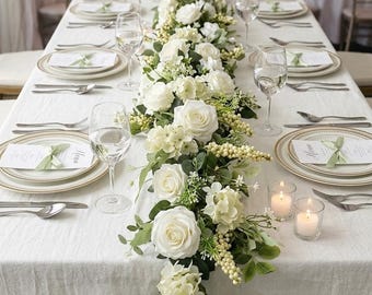 May include: A long dining table set for a formal event with a white tablecloth, white floral centerpiece featuring roses and greenery, white plates with gold rims, clear wine glasses, silver cutlery, printed menu cards with green ribbons, and candles.
