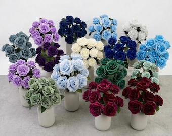 May include: A collection of artificial rose bouquets in various colors, including shades of blue, purple, red, green, white, and cream. Each bouquet is arranged in a simple, matte, light-colored cylindrical vase, displaying multiple blooming rose heads.