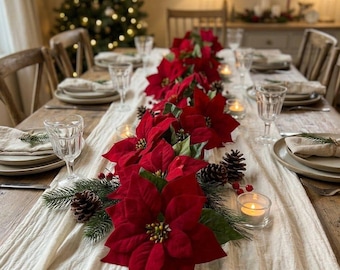 May include: A dining table set for a holiday meal featuring a long cream-colored fabric runner decorated with vibrant red poinsettia flowers, green pine branches, and brown pinecones. Small glass tea light candles are placed along the centerpiece.