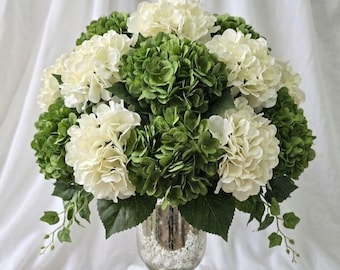 May include: A floral arrangement featuring a dense cluster of cream-white and deep green hydrangea blossoms with small green leaves, displayed in a silver-toned mercury glass pedestal vase with a textured, mottled metallic finish.
