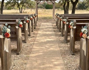 May include: An outdoor wedding aisle featuring two rows of dark wooden benches decorated with bouquets of orange, cream, and white artificial flowers tied with cream ribbons, leading to a wooden archway draped with fabric in a natural setting.