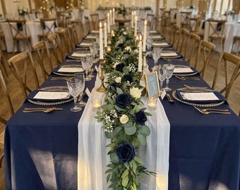 May include: A long dining table set for a formal event with a navy blue tablecloth, a white chiffon runner, and a floral centerpiece featuring dark blue roses, white roses, and eucalyptus greenery. Tall taper candles and gold table number sign.