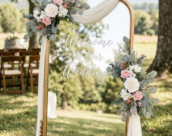 May include: A tall, rectangular gold-framed mirror featuring the white script text Welcome to Our Wedding. The frame is adorned with white and pink artificial roses, eucalyptus leaves, and draped white fabric on the top and side corners.