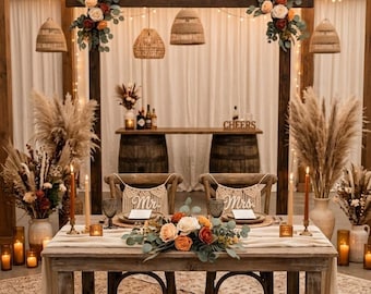 May include: A wooden sweetheart table set for two with macrame Mr. and Mrs. chair signs, floral centerpieces, taper candles, and place settings. Behind the table, a wooden pergola structure features hanging woven lights and floral accents.