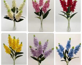May include: A collection of six artificial flower arrangements in clear glass vases, featuring tall stalks of delphinium in white, pink, red, yellow, lavender, and blue, each with green leaves and small buds at the top of the stems.