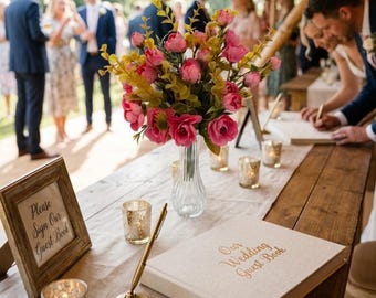 May include: A beige wedding guest book with gold text, a gold pen on a stand, a glass vase with pink flowers, and a wooden frame with the text Please Sign Our Guest Book. Several small glass votive candle holders are placed on the wooden table.
