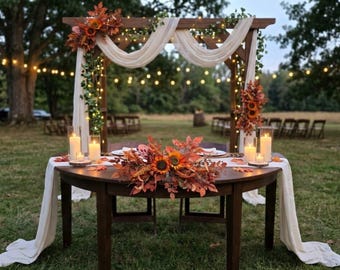 May include: A dark wood round table set for two with a white runner, autumn floral centerpiece featuring sunflowers and orange leaves, and four pillar candles in glass holders, positioned beneath a wooden arch draped in white fabric.