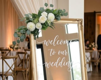 May include: A gold-framed mirror featuring the white script text "welcome to our wedding" on the glass surface. The top corner is decorated with a floral arrangement of white dahlias, white roses, and green eucalyptus leaves.