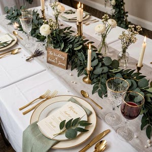 Puede incluir: Una mesa formal con mantel blanco, platos con borde dorado, cubiertos dorados y cristalería. Un centro de mesa de guirnalda de eucalipto, flores de ranúnculo blancas y velas en candelabros de latón. Un letrero de madera dice The Wedding of Sarah & James.