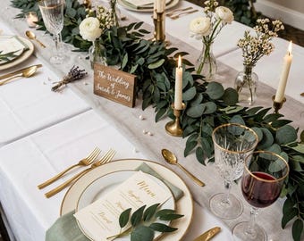 May include: A formal table setting featuring a white tablecloth, gold-rimmed plates, gold cutlery, and crystal glassware. A centerpiece of eucalyptus garland, white ranunculus flowers, and taper candles in brass holders. A wooden sign reads The Wedding of Sarah & James.