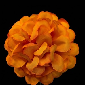 Puede incluir: Una flor artificial de color naranja vibrante con p&eacute;talos redondeados densamente superpuestos. Los p&eacute;talos de tela presentan una superficie texturizada y sutiles variaciones en tonos naranja, creando una flor esf&eacute;rica completa sobre un fondo negro s&oacute;lido.