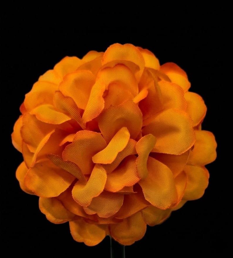 Puede incluir: Una flor artificial de color naranja vibrante con p&eacute;talos redondeados densamente superpuestos. Los p&eacute;talos de tela presentan una superficie texturizada y sutiles variaciones en tonos naranja, creando una flor esf&eacute;rica completa sobre un fondo negro s&oacute;lido.