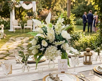 May include: A wedding table centerpiece featuring white calla lilies, roses, and hydrangeas in a glass vase. A white table card displays the text Table 7 The Roses. The setting includes gold-rimmed plates, gold cutlery, crystal glasses, and a small brass lantern.