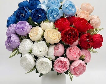 May include: A large, dense bouquet of artificial peony flowers in a wide variety of colors including deep blue, light blue, purple, white, cream, red, and pink. The flowers are arranged in a simple, solid white vase with green fern-like foliage.