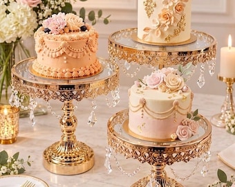 May include: Three tiered gold metal cake stands with glass tops and hanging crystal accents. Each stand holds a decorated cake featuring frosting, edible flowers, and gold detailing. The stands have ornate, filigree-style metal bases.