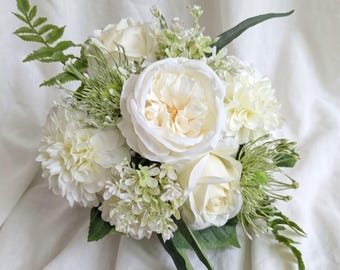 May include: A floral bouquet featuring cream-colored roses, dahlias, and small white blossoms, accented with green fern fronds and spiky green filler foliage. The arrangement is composed of various artificial flowers and greenery.