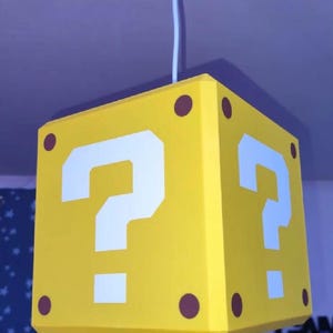 May include: A yellow cube-shaped pendant light fitting suspended from a white flex. The cube features a large white question mark on each visible side, with small brown circles positioned at each of the four corners of the square faces.