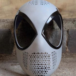 May include: A light gray full-face mask featuring large, angular eye openings outlined in glossy black trim. The mask includes a perforated pattern across the forehead and a wider perforated section covering the mouth and nose area for ventilation.