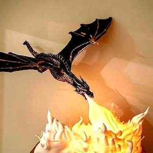 May include: A dark bronze-coloured dragon figurine in flight, positioned above a glowing, translucent white base shaped like a burst of fire. The dragon features detailed scales and outstretched wings, while the base emits a warm light.