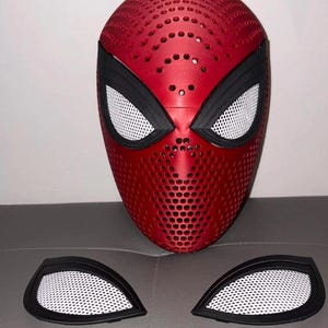 May include: A red full-face mask featuring a perforated texture throughout the surface. The mask includes black eye frames with white mesh inserts. Two additional separate black eye frames with white mesh inserts are positioned below the mask.