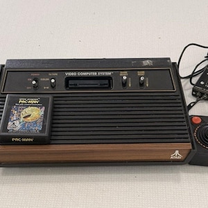 May include: An Atari Video Computer System console with a wood-grain front panel, black ribbed casing, and a Pac-Man game cartridge. The set includes two black joysticks with red buttons, two additional controllers, and various black power and signal cables.