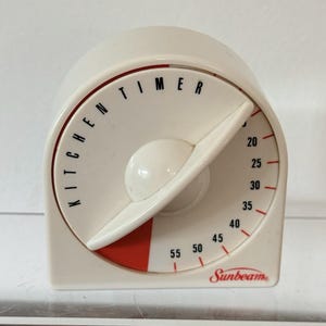 May include: A white Sunbeam kitchen timer with a rounded top and a flat base. The face features black text reading KITCHEN TIMER, black numbers from 20 to 55, and red tick marks. A large white dial pointer covers the left side with a red indicator.