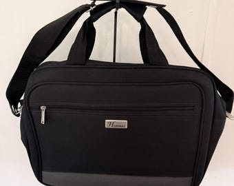 May include: A black briefcase with a horizontal zippered front pocket and a silver rectangular plaque featuring the text Hercules. The bag includes two top handles, a detachable shoulder strap, and a dark gray horizontal accent strip.