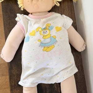 May include: A soft-sculpture doll with pale skin, blue eyes, and yellow yarn hair styled in two pigtails. The doll wears a white short-sleeved romper featuring a graphic of a yellow bunny wearing a blue dress, surrounded by small yellow hearts.