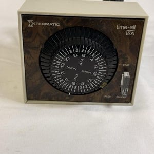 May include: An Intermatic time-all XV electrical timer with a beige plastic housing and a faux wood grain faceplate. The device features a large black circular dial with white printed numbers, AM and PM indicators, and a clear plastic on-off switch.