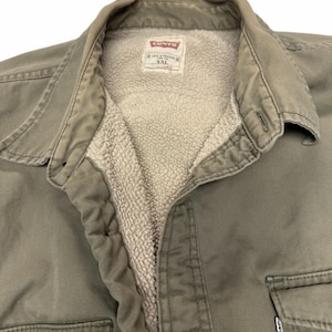 May include: An olive green Levi's jacket with a beige sherpa lining. The collar is folded, and the interior label displays the Levi Strauss and Co. logo with the size XXL. A small white Levi's brand tag is visible on the front chest pocket.