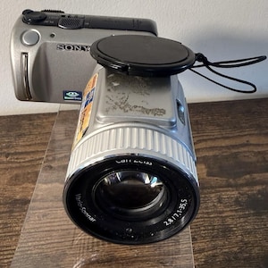 May include: A silver Sony digital camera featuring a black Carl Zeiss Vario-Sonnar 2.8/7.1-35.5 lens. The camera body has a black lens cap attached by a cord, a Memory Stick logo, and a rectangular sticker on the top surface near the lens barrel.