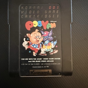 May include: A black video game cartridge for the Atari Video Game System and Sears Video Arcade. The label features the title Pooyan, a pig and a wolf character, and the text Konami 001 Video Game Cartridge, Konami Industry Co. Ltd 1982.