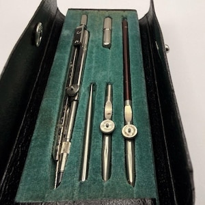 May include: A vintage drafting instrument set housed in a black leather case with a teal velvet interior. The set includes a large metal compass, a small metal needle point, two smaller metal compass attachments with adjustment wheels, and a lead holder.