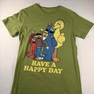 May include: An olive green short-sleeve t-shirt featuring a graphic print of Sesame Street characters Elmo, Ernie, Bert, Cookie Monster, and Big Bird. Below the characters, the text "HAVE A HAPPY DAY" is printed in yellow capital letters.
