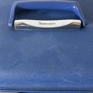 May include: A blue hardshell suitcase with a textured exterior finish. The top features a blue plastic handle attached to a silver-toned metal plate engraved with the brand name Samsonite. The case includes silver-toned metal latches along the bottom edge.
