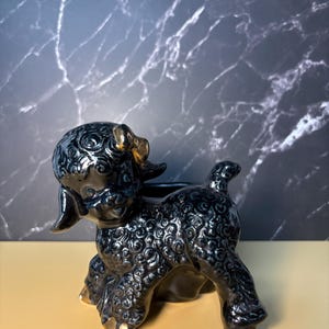 May include: A glossy black ceramic poodle figurine featuring textured curly fur, a gold-painted bow on its head, and gold-painted accents on its paws. The poodle is standing in a side profile position with its head turned toward the front.