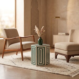 May include: A hexagonal side table featuring a white and dark green geometric pattern, positioned between two beige upholstered chairs. A dark green vase containing dried grasses sits on the table surface, which has a smooth, flat top.