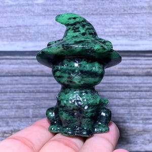May include: A small, carved stone figurine of a cat wearing a pointed witch hat. The sculpture is made of a dark green mineral with black speckled inclusions throughout the surface, featuring a smooth, polished texture and a seated posture.