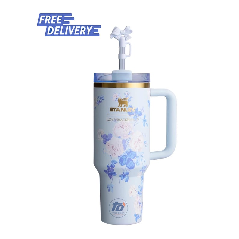 May include: A light blue insulated tumbler with a floral pattern, a gold rim, and a side handle. The Stanley and LoveShackFancy logos are printed on the front. A clear lid features a straw topped with a white bow accessory. The base displays a logo.