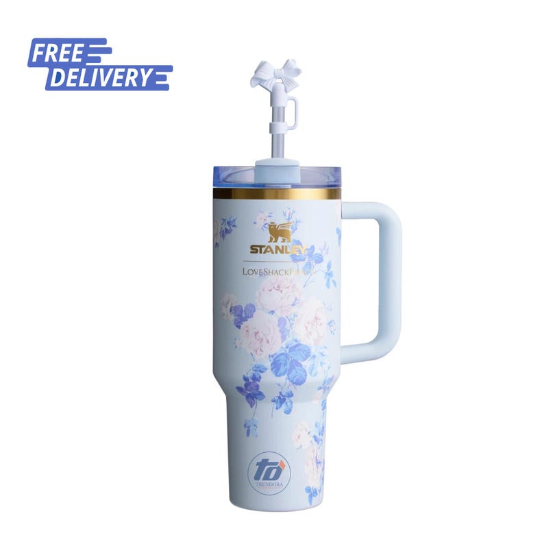 May include: A light blue insulated tumbler with a handle, featuring a floral pattern in shades of blue and pink. The cup displays the Stanley and LoveShackFancy logos, a gold rim, a clear lid, and a straw topped with a white bow accessory.
