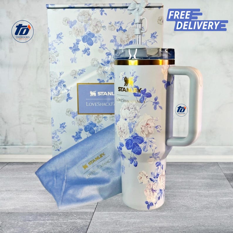 May include: A white stainless steel tumbler with a blue floral pattern, featuring the Stanley and LoveShackFancy logos, a clear lid, a light blue handle, and a matching floral box with a blue fabric pouch displaying the Stanley logo.