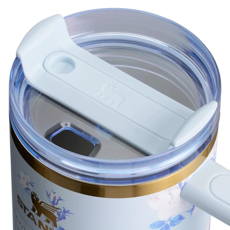 May include: A light blue tumbler lid with a clear plastic rim and a sliding closure mechanism. The lid features a light blue sliding tab embossed with a winged bear logo. A gold-colored metal ring encircles the top edge of the tumbler body.